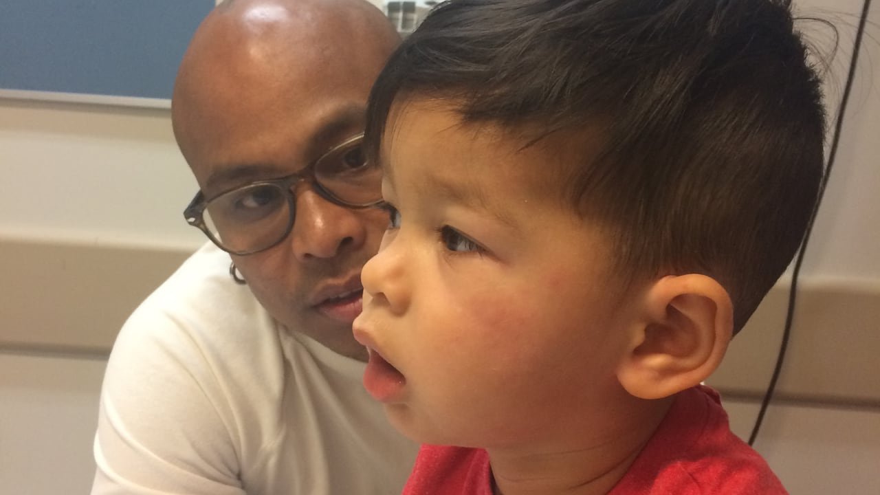 default-160 In a hospital room, a man looks concerned at a boy breathing with his mouth open and who has hives on his cheeks.
