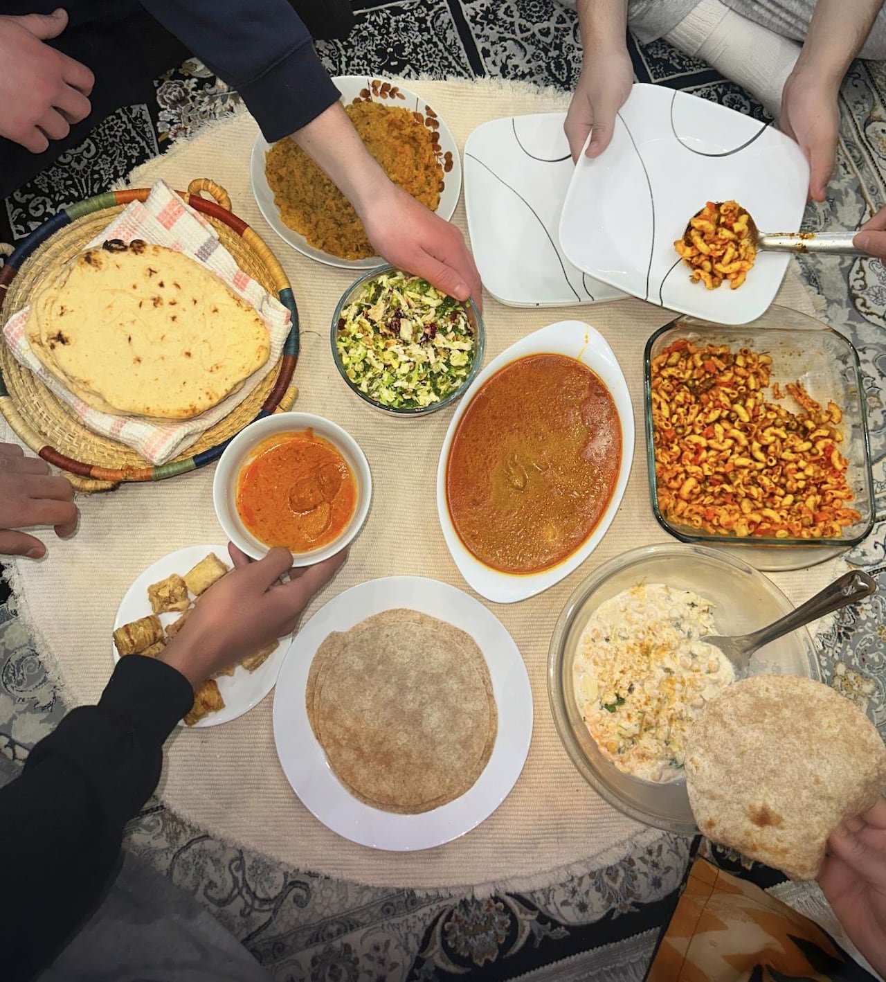 default-230 A bird’s eye view of several hands reaching for food, such as butter chicken, macaroni salad, roti and baklava.