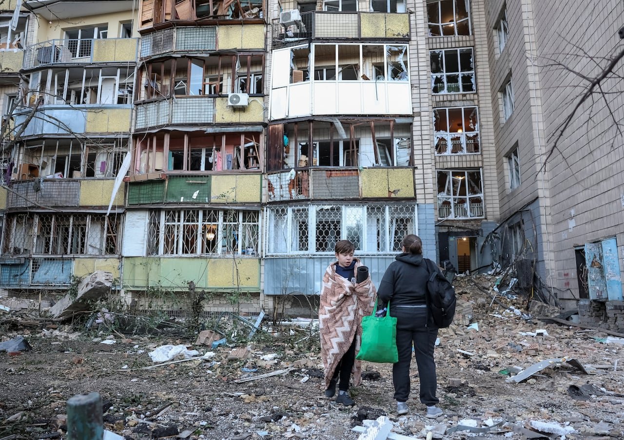 A boy wrapped in a blanket stands outside a damaged apartment building beside another resident.
