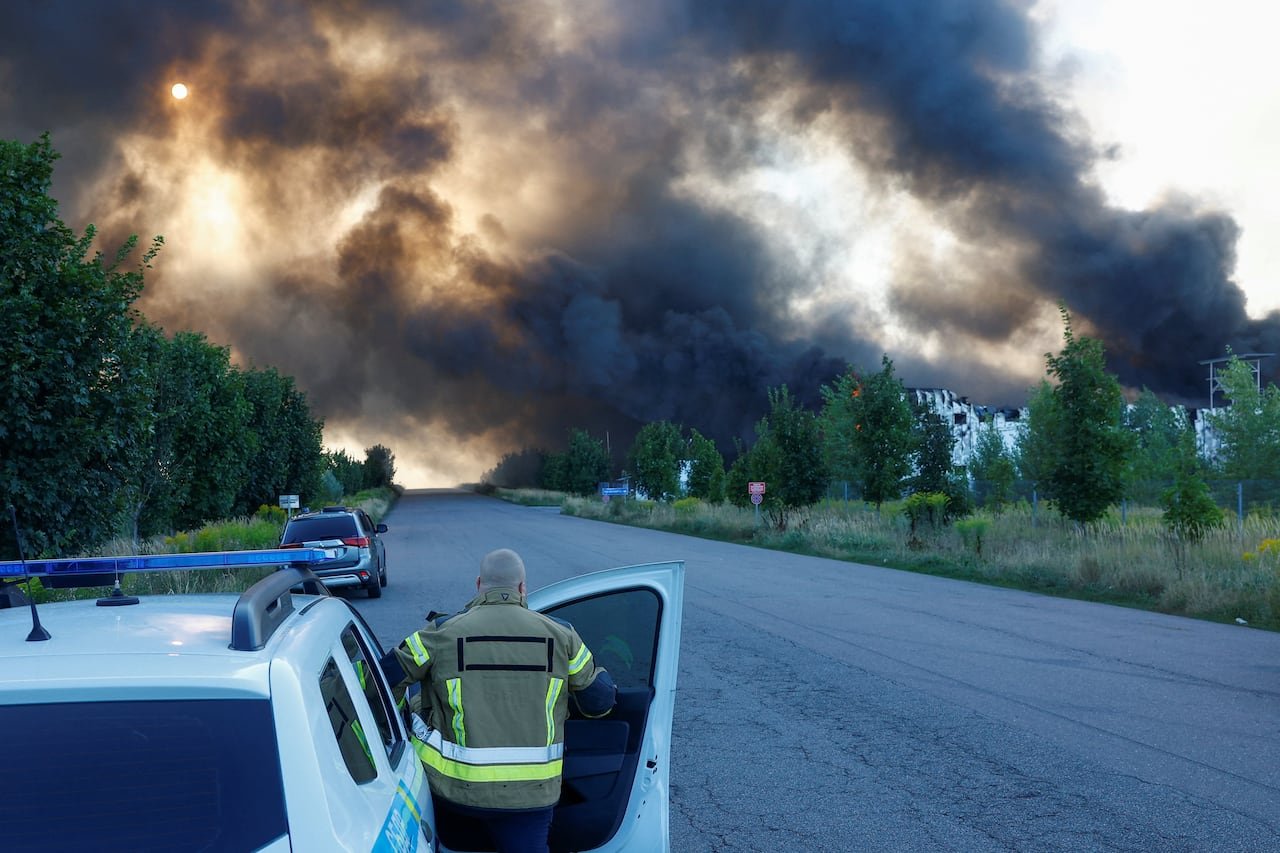 A firefighter with his back to the camera gets out of the passenger side of a vehicle as off in the distance of a rural area, large black clouds of smoke fill the sky near a series of low-rise structures.