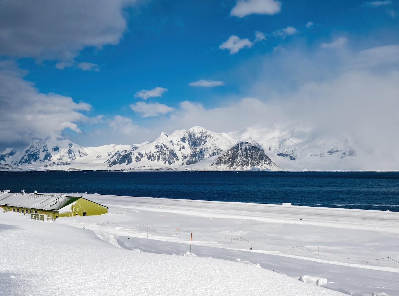 A low green building, half buried in snow, next to a lake with snowy mountains on the horizon