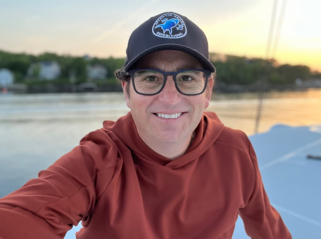 default-542 A smiling man with square glasses. He is wearing a baseball hat and an orange hoodie sitting on a boat.